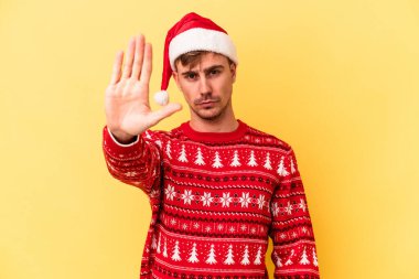 Genç, beyaz bir adam, sarı arka planda izole edilmiş bir şekilde Noel 'i kutluyor. Elini uzatmış, dur işareti gösteriyor..