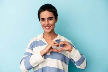 Mavi arka planda izole edilmiş genç beyaz kadın gülümsüyor ve elleriyle kalp şeklini gösteriyor..