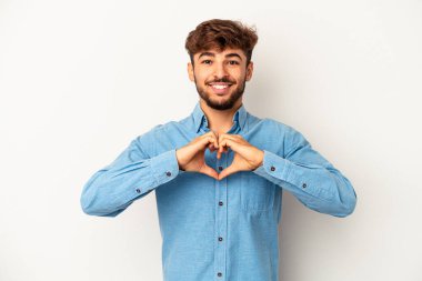 Genç melez adam gri arka planda izole edilmiş gülümsüyor ve elleriyle kalp şeklini gösteriyor..