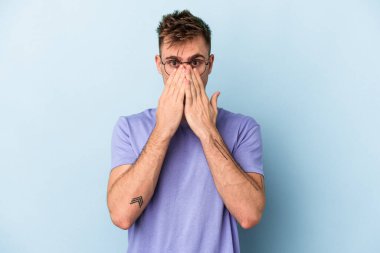 Mavi arka planda izole edilmiş genç beyaz adam şok olmuş, ağzı ellerle kaplanmış, yeni bir şey keşfetmeye hevesli..