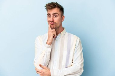 Mavi arka planda izole edilmiş genç beyaz adam alaycı bir ifadeyle kameraya bakarken mutsuz oluyor..