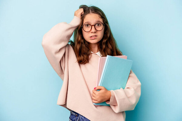 Little caucasian student girl holding books isolated on blue background being shocked, she has remembered important meeting.