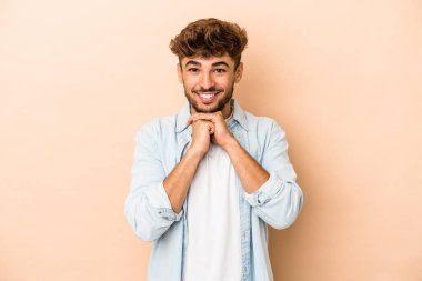 Genç Arap adam bej arka planda izole edilmiş ellerini çenesinin altında tutuyor, mutlu bir şekilde yana bakıyor..