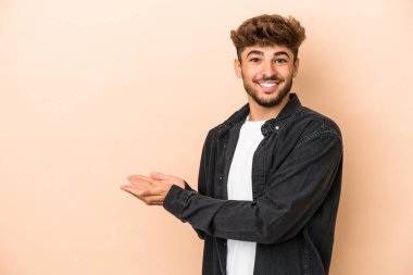 Young arab man isolated on beige background holding a copy space on a palm.