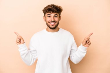 Young arab man isolated on beige background pointing to different copy spaces, choosing one of them, showing with finger.