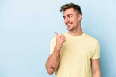 Kafkasyalı genç adam mavi arka plan noktalarında izole edilmiş başparmak uzakta, gülüyor ve kaygısız.