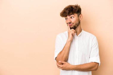 Genç Arap adam bej arka planda izole edilmiş. Şüpheli ve şüpheci bir ifadeyle yana bakıyor..