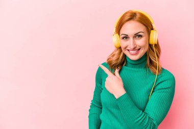 Pembe arka planda izole edilmiş müzik dinleyen beyaz bir kadın gülümsüyor ve boş alanda bir şeyler gösteriyor..