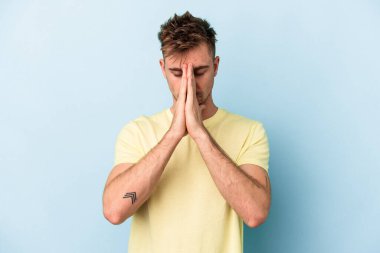 Mavi arka planda izole edilmiş genç beyaz bir adam el ele tutuşup, ağzının kenarında dua ediyor, kendinden emin..