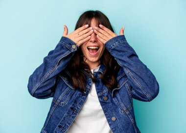 Mavi arka planda izole edilmiş genç Arjantinli kadın elleriyle gözlerini kapatıyor, geniş bir gülümsemeyle bir sürpriz bekliyor..