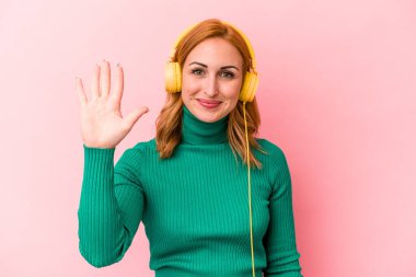 Pembe arka planda izole edilmiş müzik dinleyen beyaz bir kadın gülümseyerek beş numarayı parmaklarıyla gösteriyor..