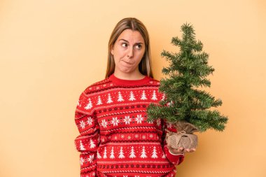 Sarı arka planda izole edilmiş küçük bir noel ağacı tutan genç beyaz kadın şaşkın ve kararsız hissediyor..