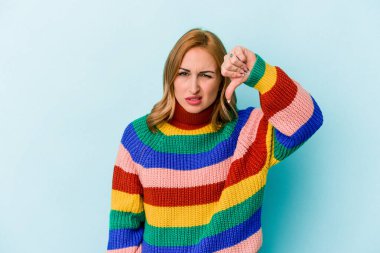 Mavi arka planda izole edilmiş genç beyaz kadın baş parmağını aşağı gösteriyor ve hoşlanmadığını ifade ediyor..