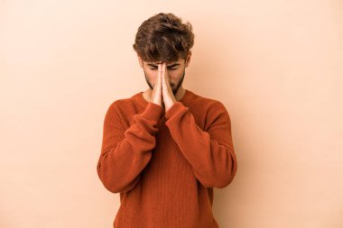 Young arab man isolated on beige background holding hands in pray near mouth, feels confident.