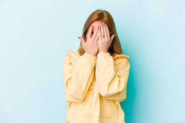 Mavi arka planda izole edilmiş genç beyaz kadın kameraya parmaklarıyla göz kırpıyor, yüzünü kapatmaktan utanıyor..