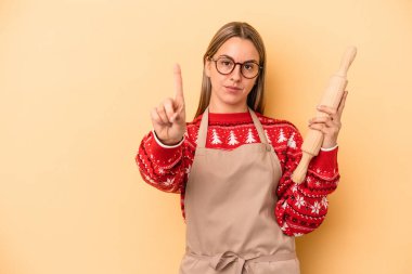 Genç fırıncı kadın Noel için kurabiye yapıyor. Sarı arka planda izole edilmiş. Parmak izleriyle bir numarayı gösteriyor..