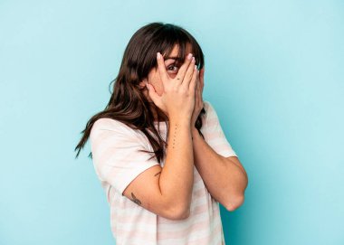 Arjantinli genç bir kadın mavi arka planda izole edilmiş. Korkmuş ve gergin parmaklarıyla göz kırp..