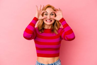 Pembe arka planda izole edilmiş genç beyaz kadın bir başarı fırsatı bulmak için gözlerini açık tutuyor..