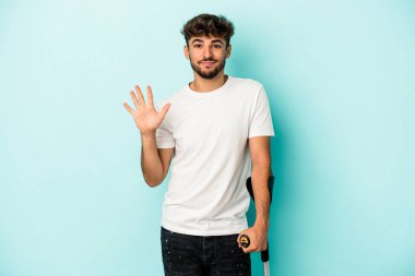 Young arab man with crutches isolated on blue background smiling cheerful showing number five with fingers.