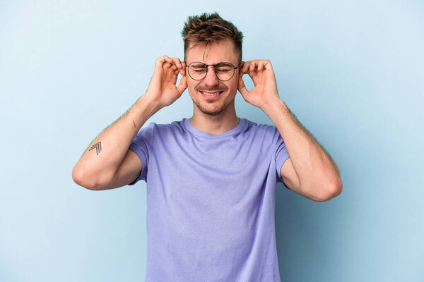 Молодой кавказский мужчина изолирован на синем фоне, закрывая уши руками.