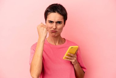 Elinde cep telefonu olan beyaz bir kadın pembe arka planda izole edilmiş, yumruğunu kameraya gösteriyor, agresif yüz ifadesi var..