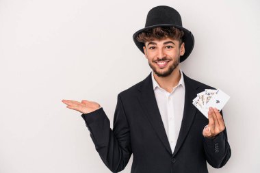 Young arab wizard man holding a magic cards isolated on white background showing a copy space on a palm and holding another hand on waist.