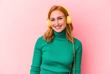 Pembe arka planda izole edilmiş müzik dinleyen genç beyaz kadın mutlu, güler yüzlü ve neşeli..