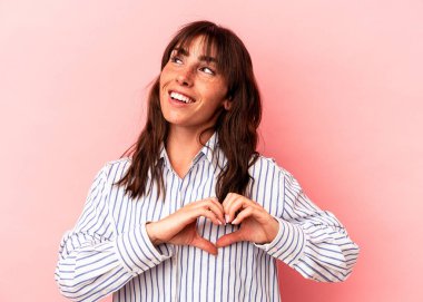 Pembe arka planda izole edilmiş genç Arjantinli kadın gülümsüyor ve elleriyle kalp şeklini gösteriyor..