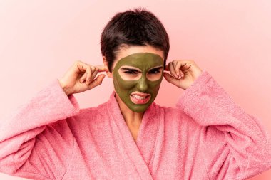 Yüz maskesi takan beyaz bir kadın pembe arka planda izole edilmiş kulakları elleriyle kapatıyor..