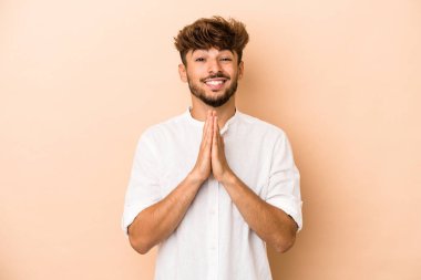 Young arab man isolated on beige background holding hands in pray near mouth, feels confident.