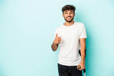 Young arab man with crutches isolated on blue background smiling and raising thumb up