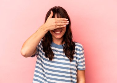 Pembe arka planda izole edilmiş genç Arjantinli kadın elleriyle gözlerini kapatıyor, geniş bir gülümsemeyle bir sürpriz bekliyor..