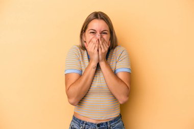 Sarı arka planda izole edilmiş genç beyaz kadın bir şeye gülüyor, elleriyle ağzını kapatıyor..