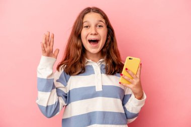 Pembe arka planda cep telefonu tutan küçük beyaz kız hoş bir sürpriz alıyor, heyecanlı ve el kaldırıyor..