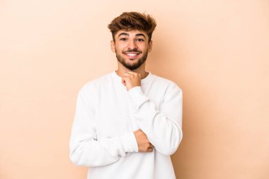 Young arab man isolated on beige background smiling happy and confident, touching chin with hand.