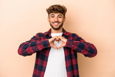 Genç Arap adam bej arka planda izole edilmiş gülümsüyor ve elleriyle kalp şekli gösteriyor..