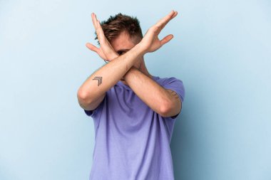 Kafkasyalı genç adam mavi arka planda izole edilmiş iki kolu çapraz tutuyor, inkar kavramı.