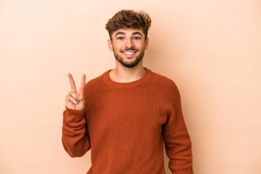 Genç Arap adam bej arka planda izole edilmiş zafer işareti gösteriyor ve gülümsüyor..
