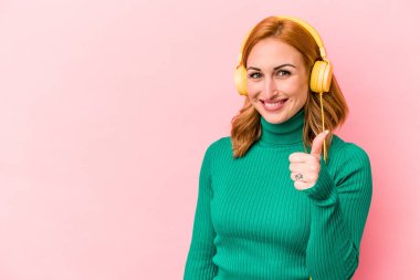 Pembe arka planda izole edilmiş müzik dinleyen beyaz bir kadın gülümsüyor ve başparmağını kaldırıyor.