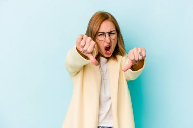 Mavi arka planda izole edilmiş genç beyaz kadın baş parmağını aşağı gösteriyor ve hoşlanmadığını ifade ediyor..