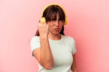 Arjantinli genç bir kadın pembe arka planda izole edilmiş müzik dinliyor. Kameraya yumruk, agresif yüz ifadesi gösteriyor..