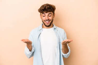 Young arab man isolated on beige background holding something with palms, offering to camera.