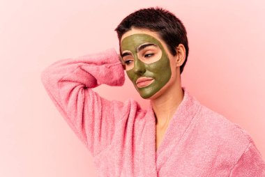 Yüz maskesi takan beyaz bir kadın pembe arka planda izole edilmiş kafasının arkasına dokunuyor, düşünüyor ve bir seçim yapıyor..