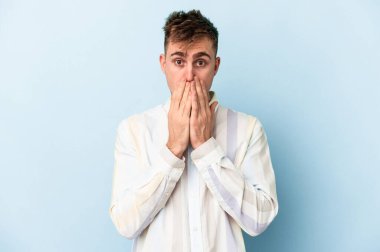 Mavi arka planda izole edilmiş genç beyaz adam elleriyle ağzını kapatırken şok oldu..