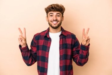 Genç Arap adam bej arka planda izole edilmiş zafer işareti gösteriyor ve gülümsüyor..