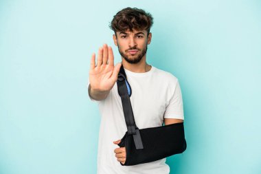 Kırık elli genç bir Arap, mavi arka planda izole edilmiş, elini uzatmış, dur işareti gösteriyor, bu da seni engelliyor..