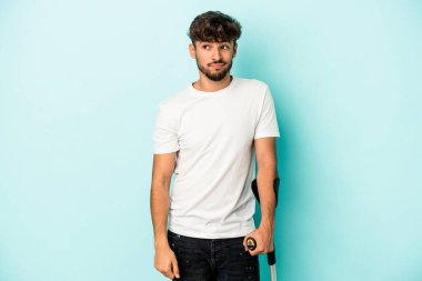 Koltuk değnekli genç Arap adam mavi arka planda izole edilmiş, kafası karışmış, şüpheli ve kendinden emin değil..