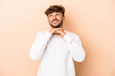 Genç Arap adam bej arka planda izole edilmiş ellerini çenesinin altında tutuyor, mutlu bir şekilde yana bakıyor..