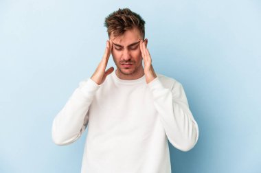 Genç beyaz adam mavi arka planda izole edilmiş tapınaklara dokunuyor ve başı ağrıyor..