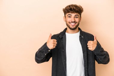 Genç Arap adam bej arka planda izole edilmiş gülümsüyor ve başparmağını kaldırıyor.